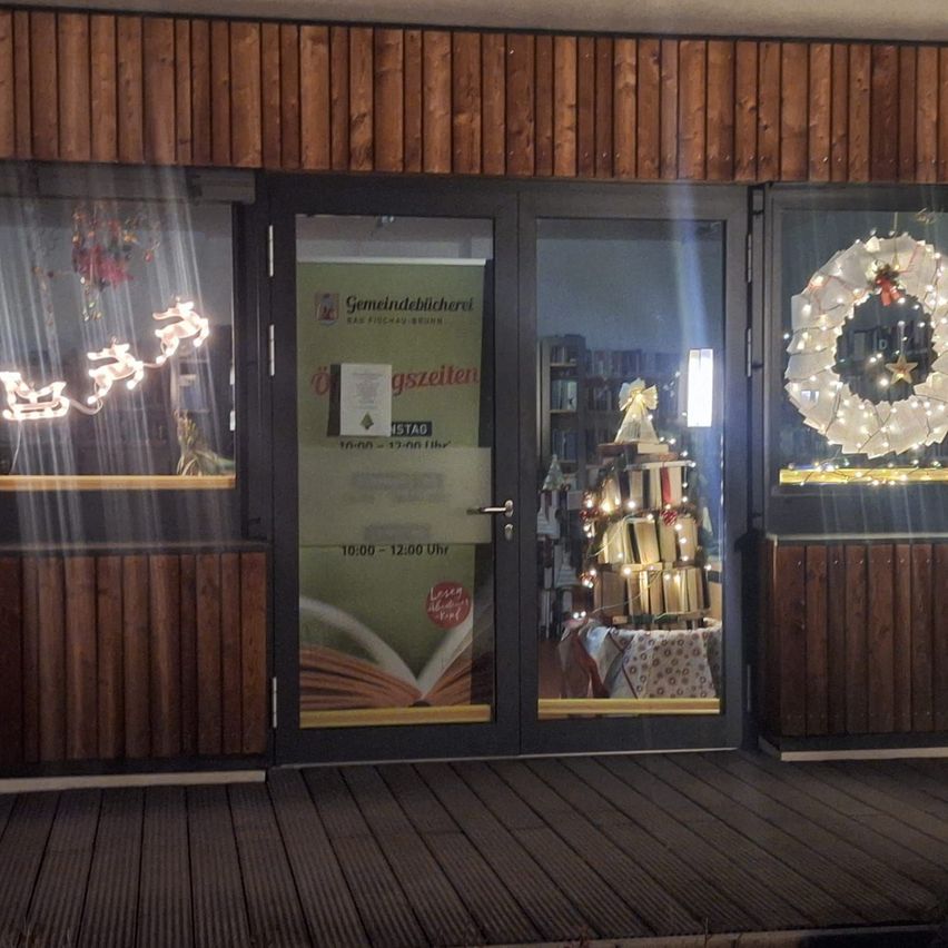 Ein hölzerner Gebäudeeingang mit Glastüren zeigt eine beleuchtete Weihnachtsgirlande und eine Engelsfigur. Ein Schild mit Öffnungszeiten und einem offenen Buch deutet auf die Öffnungszeiten hin, und Lichter reflektieren sich auf der Scheibe.