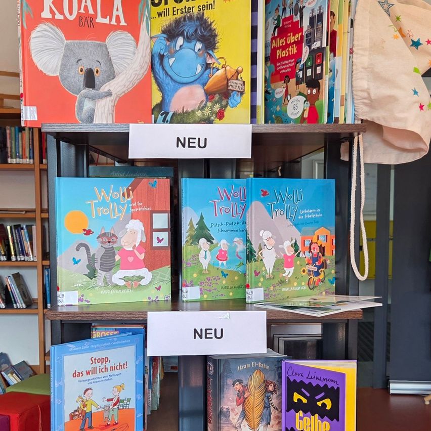 Ein Holzregal zeigt mehrere Kinderbücher, darunter 'Koala Bar' und die 'Wolli Trolly'-Reihe. Ein weißes Schild mit 'NEU' ist über den Büchern angebracht.