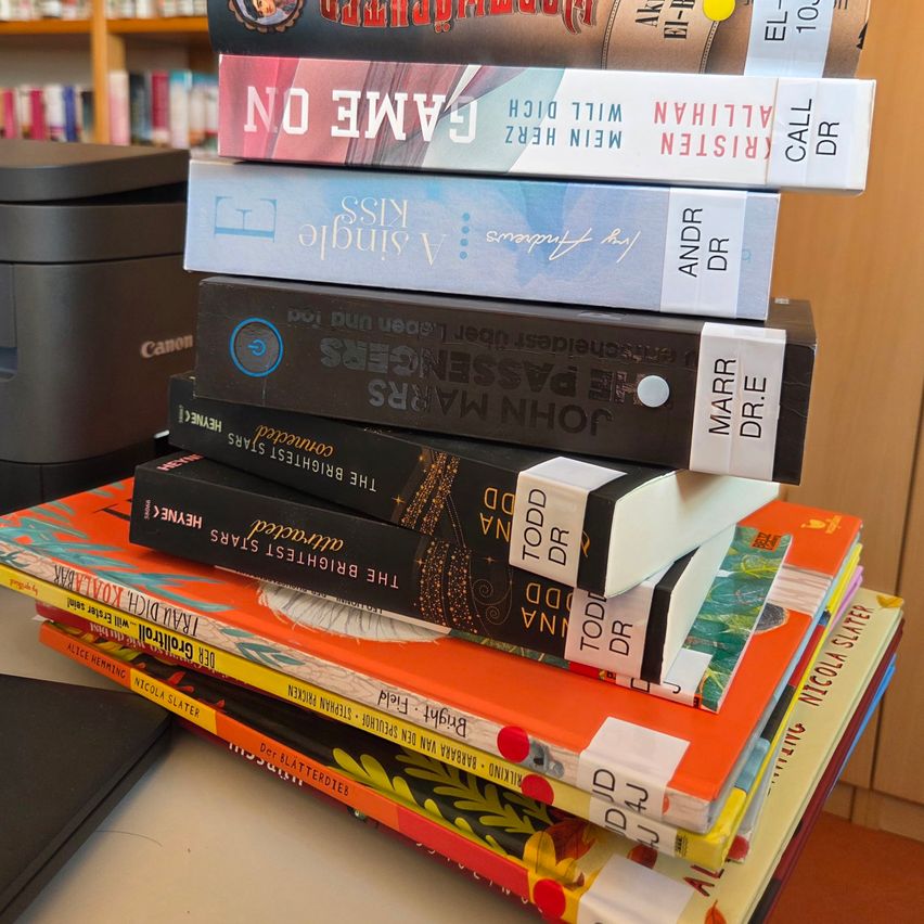 Ein Stapel Bücher mit Titeln wie 'Die verschwundene Hälfte', 'Die stille Patientin' und 'Der Nachtzirkus' sind in einer Bibliothek auf einem Tisch angeordnet.