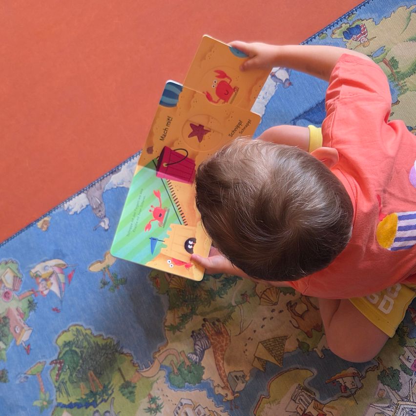 Ein Baby sitzt auf einem farbenfrohen Teppich und schaut ein Buch an, auf dessen Cover ein Krabbe abgebildet ist.