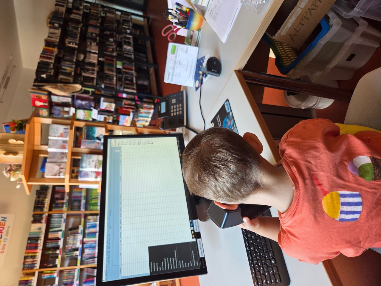 Ein junger Junge in einem orangefarbenen T-Shirt sitzt in einer Bibliothek vor einem Computer, hält ein Telefon an sein Ohr und schaut auf den Monitor.