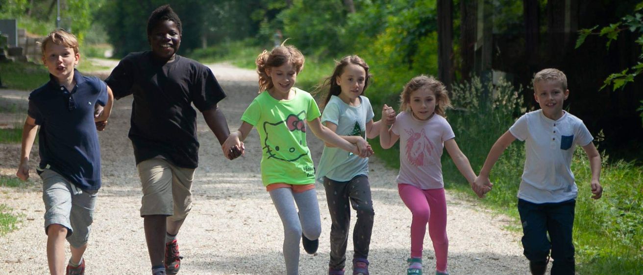 Mehrere Kinder halten Händchen und laufen auf einem Kiesweg, umgeben von Bäumen.