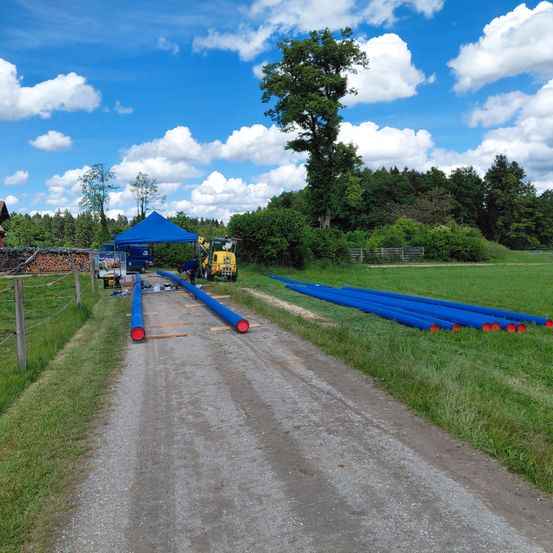 Eine ländliche Straße mit blauen Rohren, die auf der Seite verlegt sind, einem blauen Zelt, einem gelben Maschinenfahrzeug und einem Haus in der Ferne unter einem blauen Himmel mit Wolken.