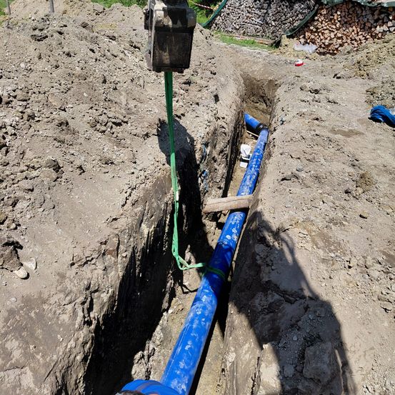 Eine blaue Rohrleitung wird in einer Grube verlegt, wobei ein mechanischer Arm sie anhebt. Die Grube ist mit Erde und Steinen gefüllt, und in der Nähe befinden sich Holzstapel.