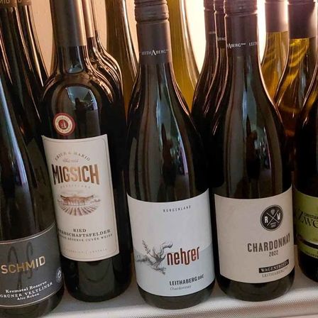Eine Reihe von Weinflaschen auf einem Regal, darunter Migisch, Schmid, Neber und Chardonnay. Die Neber-Flasche ist mit Leithaberg DAC Chardonnay beschriftet.