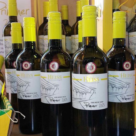 Eine Reihe von dunklen Flaschen mit gelben Verschlüssen, beschriftet mit 'Heiss Primaner 2023', stehen in einem Geschäft. Die Flaschen stehen eng beieinander.