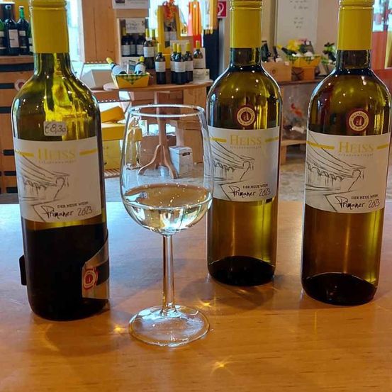 Drei Flaschen Weißwein mit dem Etikett 'Heiss' stehen auf einem Tisch mit einem Weinglas. Die Weinflaschen haben Etiketten, die das Jahr 2023 anzeigen. Im Hintergrund befinden sich Regale mit weiteren Flaschen und verschiedenen Artikeln.