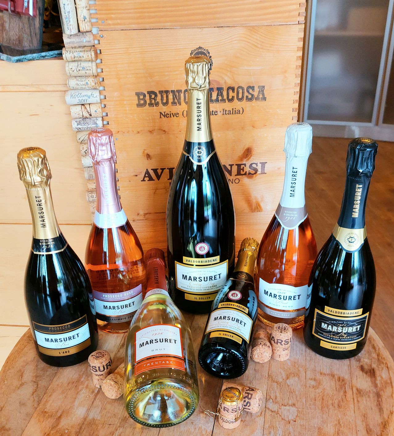 Eine Sammlung von Prosecco- und Marsuret-Flaschen, die auf einer Holzoberfläche mit Korken und einer Holzkiste mit der Aufschrift Bruno Lacosta im Hintergrund angeordnet sind.