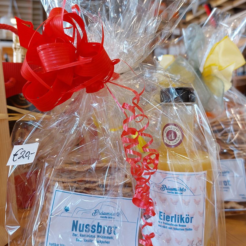 Ein Geschenkkorb mit rotem Band enthält eine Flasche 'Eierlikör' und 'Nussbrot'. Die Artikel sind in durchsichtiger Plastikfolie verpackt. Das Preisschild zeigt 'E20'. Im Hintergrund sind weitere Geschenkkörbe und Regale mit Produkten zu sehen.