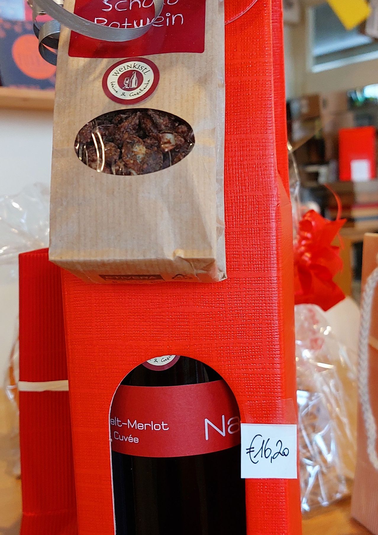 Eine rote Box mit einem durchsichtigen Fenster zeigt eine Weinflasche mit dem Etikett 'N3 elt-Merlot Cuvee'. Ein brauner Beutel mit einem Logo ist obenauf, und ein roter Bogen schmückt die Seite.