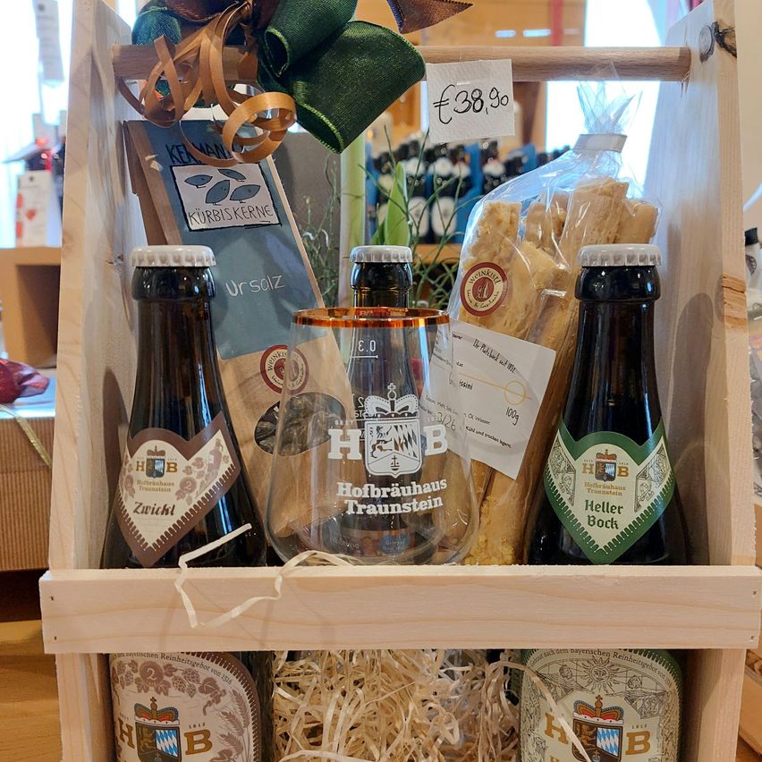 Eine Holzkiste enthält ein Set mit drei Flaschen, einem Glas und einer Lebensmittelpackung. Die Flaschen sind mit Hofbrauhaus Traunstein beschriftet. Die Kiste hat einen Preisschild von 38,90 Euro.