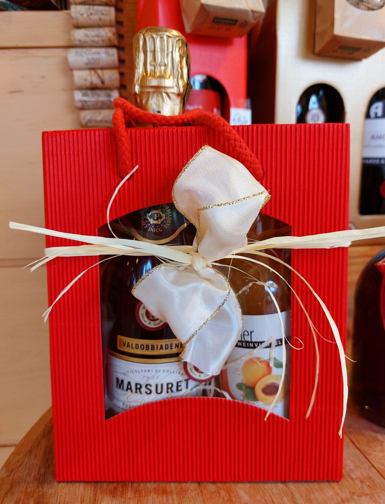 Ein rotes Geschenktüte mit einer Schleife enthält zwei Flaschen. Eine ist mit 'Marsuret' und die andere mit 'Weinverze' beschriftet.