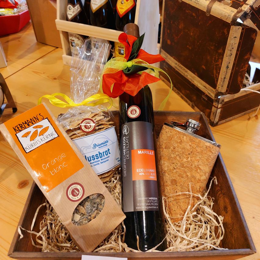 Eine Holzkiste enthält eine Flasche Marille-Wein, einen Flakon und eine Tüte orangefarbener Snacks, mit einem dekorativen Band und Blatt obenauf.