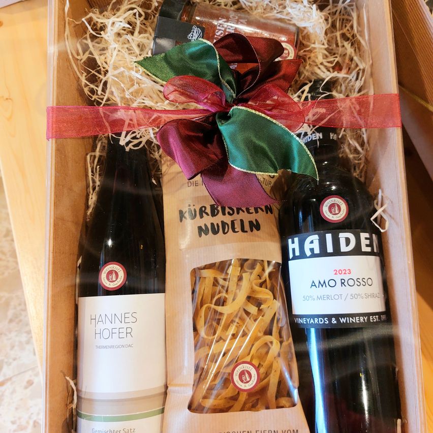 Eine Holzkiste enthält zwei Flaschen Wein und einen Beutel mit Nudeln, verziert mit einer roten und grünen Schleife obenauf.