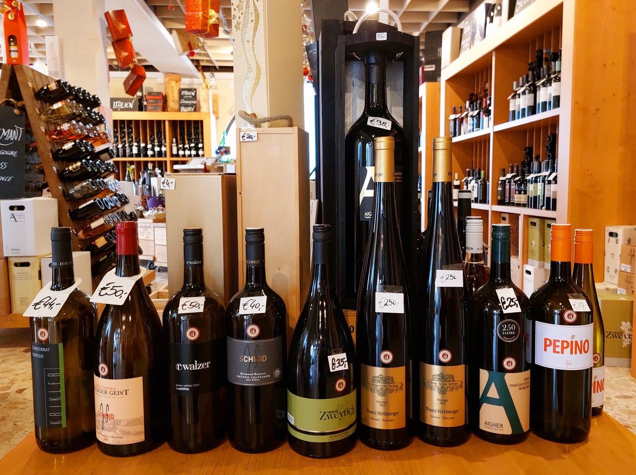 Verschiedene Flaschen Wein sind in einem Geschäft ausgestellt, darunter eine mit dem Etikett 'Aigner'. Jede Flasche hat einen Preisschild, wobei die Preise von 25 bis 50 Euro reichen.