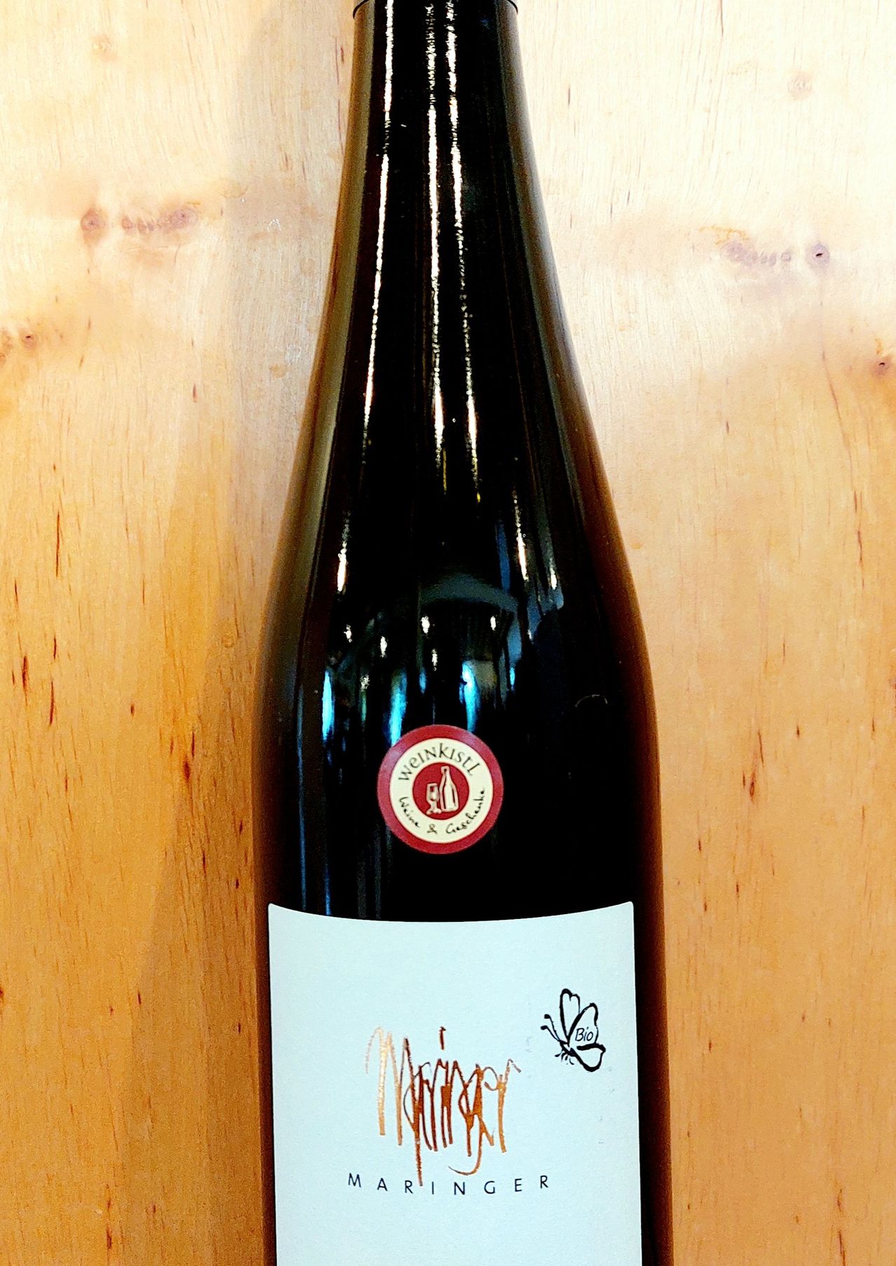 Eine Flasche Rotwein mit einem roten kreisförmigen Aufkleber mit Logo, auf einem holzfarbenen Hintergrund. Das Etikett hat handgeschriebenen Text und eine Schmetterlingsillustration.