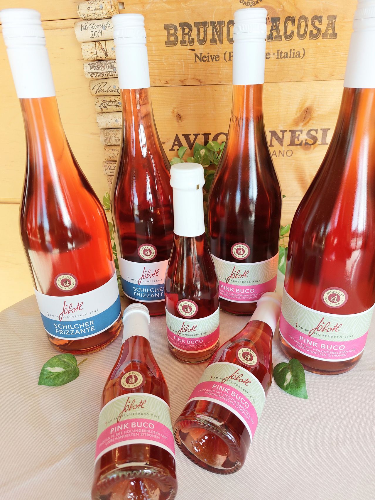 Five bottles of pink bubbly wine with white caps sit on a table. Three bottles are larger, two are smaller. The table is covered with a white cloth. Behind the bottles, there is a wooden box with text.