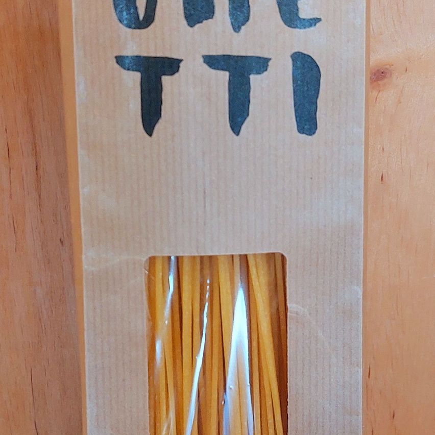 Ein Nahaufnahme eines braunen Kartonpakets mit blauem Text, der 'UNL TTI' liest. Im Inneren des Pakets befindet sich ein klares Kunststofffenster, das einen Stapel gelber Nudeln zeigt.
