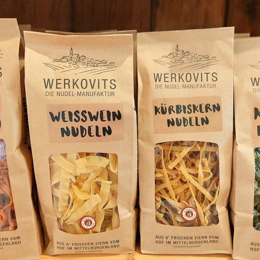 Vier Tüten mit Nudeln sind auf einem Holzregal ausgestellt. Die Tüten sind braun und haben die Aufschrift 'Werkovit's Die Nudel-Manufaktur' und 'Weisswein Nudeln' und 'Kürbiskern Nudeln'. Der Inhalt der Nudeln ist durch das transparente Fenster sichtbar.