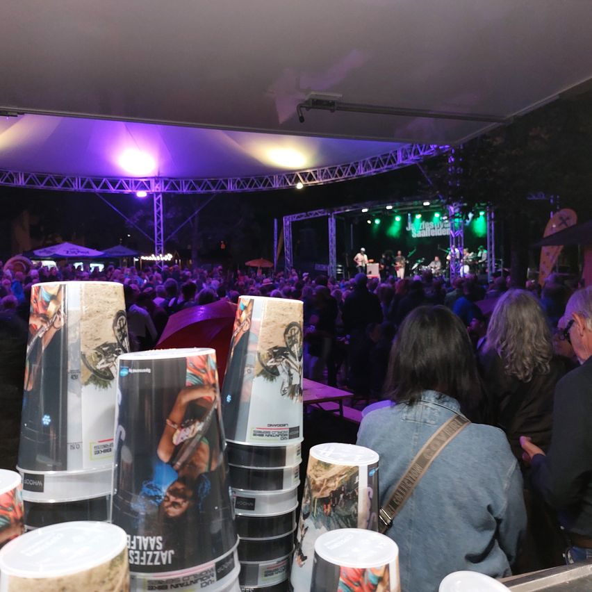 Ein nächtliches Open-Air-Konzert mit einer großen Menschenmenge, einer Bühne mit einer Band, die unter lila Beleuchtung auftritt, und Bechern mit Bildern von Menschen darauf.