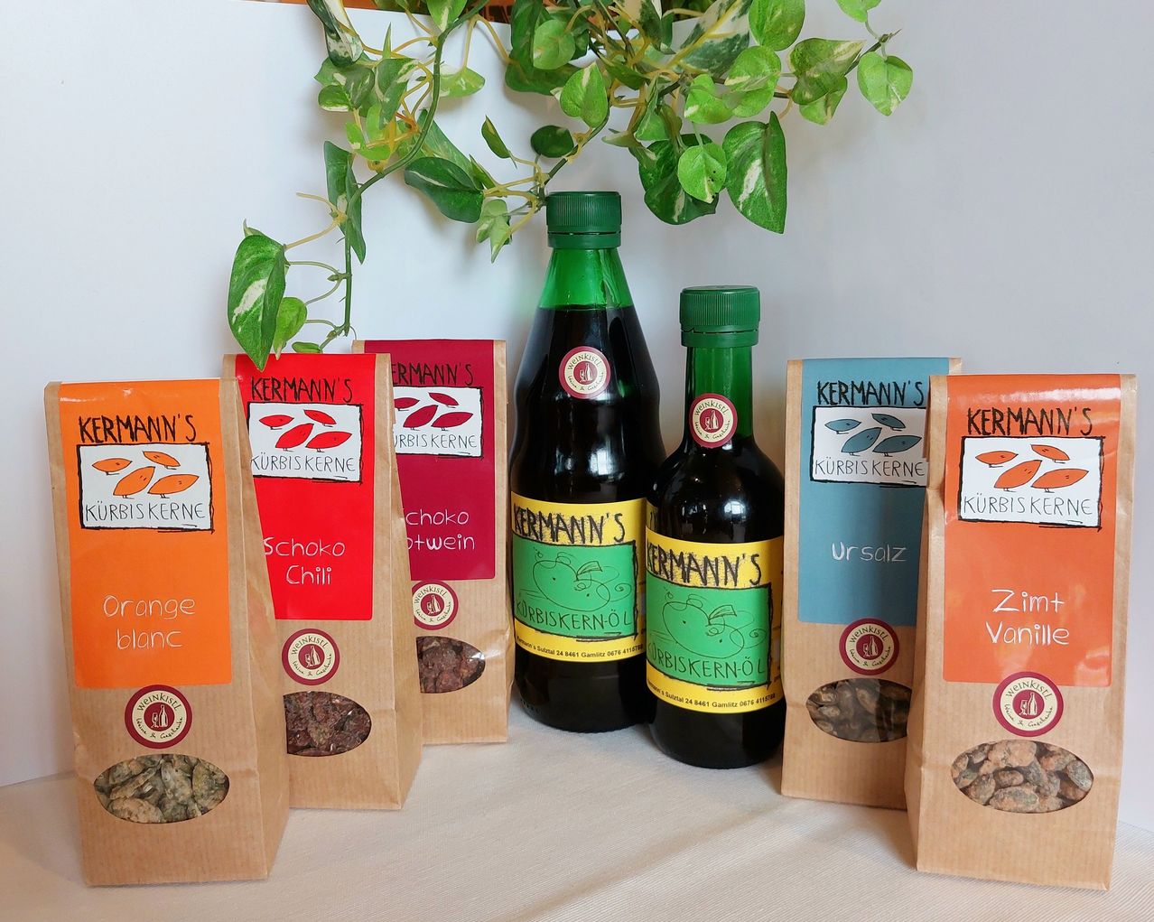 Eine Ausstellung von Kermann-Produkten, darunter zwei Flaschen Kürbiskernöl, eine mit grünem Etikett und die andere mit gelbem Etikett, und vier Beutel Kürbiskerne in verschiedenen Geschmacksrichtungen.