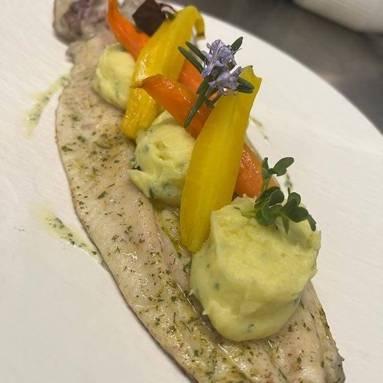 Ein weißer Teller mit einem Fischfilet, belegt mit Kartoffelpüree, gelben und orangefarbenen Paprikaschoten und Kräutern. Das Gericht ist mit einer violetten Blume garniert.