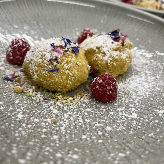 Zwei goldbraune Gebäckstücke, bestreut mit Puderzucker, garniert mit frischen Himbeeren und blauen essbaren Blüten, präsentiert auf einem grauen Teller.