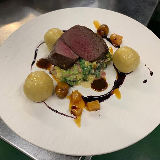 Ein weißer Teller mit einem geschnittenen Rindersteak, Kartoffelpüree und grünem Gemüse. Das Gericht ist mit einer Saucensoße und kleinen Fruchtstücken garniert.
