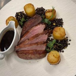 Ein weißer Teller mit geschnittenem Fleisch, kleinen Kartoffeln, schwarzen Bohnen und einem kleinen Becher Sauce.