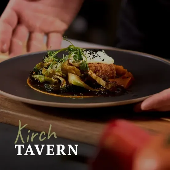 Die Hand eines Kochs, die einen Teller mit einem Gericht aus Brokkoli, Fleisch und Sauce hält, mit den Worten 'Kirch Tavern' im Vordergrund.
