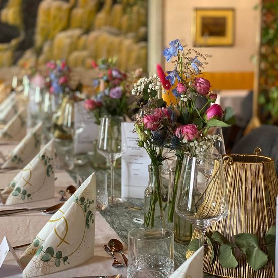 Ein langer Tisch für eine besondere Veranstaltung mit Blumensträußen als Mittelpunkt, Weingläsern und dekorativen Servietten. Der Tisch ist mit verschiedenen Blumen und Grünpflanzen geschmückt.