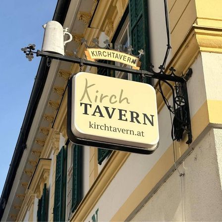 Ein gelbes Schild mit den Worten 'Kirch Tavern' hängt an einem Gebäude mit grünen Fensterläden und einer weißen Keramiktasse oben.