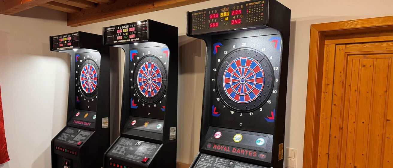 Drei Arcade-Dartautomaten sind an einer Wand aufgestellt, mit Dartscheiben und digitalen Anzeigetafeln, einer mit der Aufschrift 'Royal Darts'.