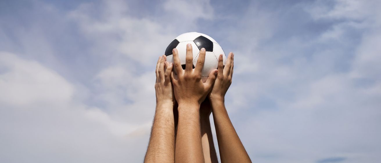 Mehrere Hände halten einen Fußball unter einem blauen Himmel mit Wolken. Der Ball ist weiß und schwarz, mit schwarzen Mustern.