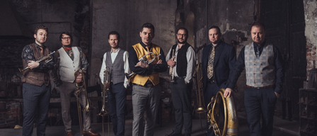 Eine Gruppe von Musikern steht in einem schwach beleuchteten Raum mit alten Wänden. Sie halten Blechblasinstrumente.