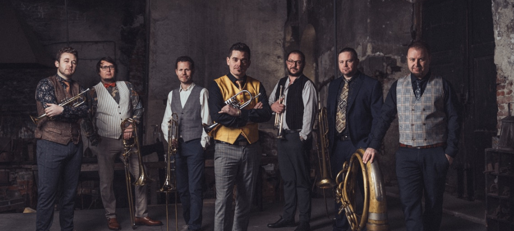 Eine Gruppe von Musikern steht in einem schwach beleuchteten Raum mit alten Wänden. Sie halten Blechblasinstrumente.