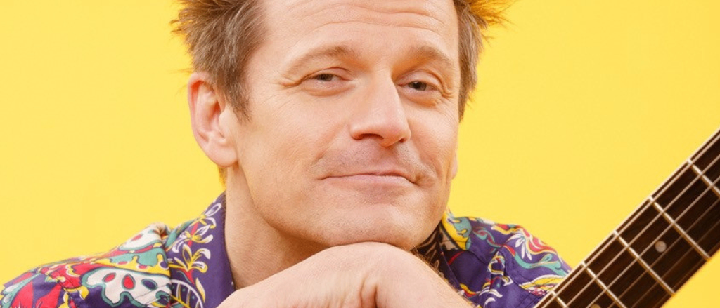 Ein Mann mit struppigem Haar lächelt und posiert für ein Foto, während er eine Gitarre hält. Er trägt ein farbenfrohes Hemd mit einem Design.
