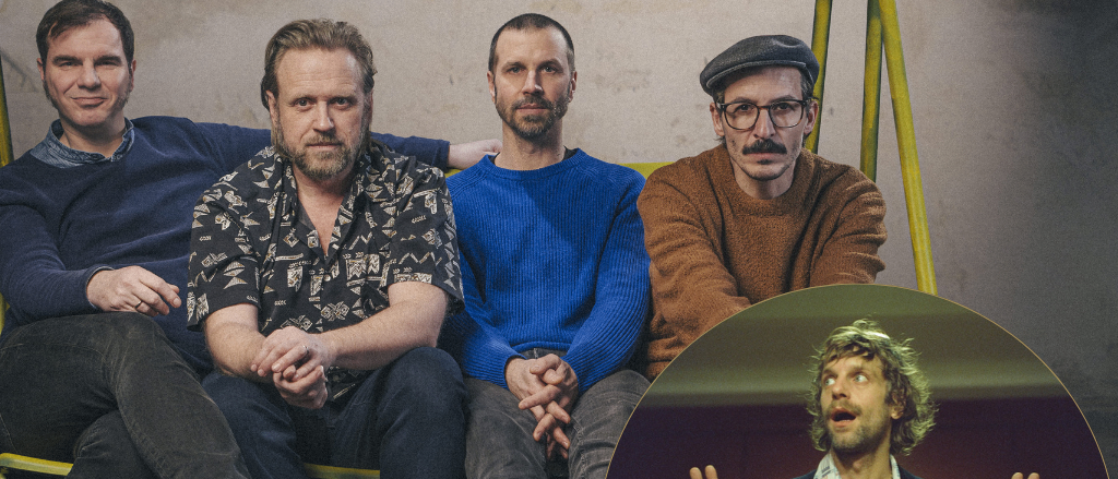 Vier Männer sitzen auf einer Bank. Einer trägt ein Blumenhemd, ein anderer ein blaues Sweatshirt, während der Dritte ein braunes Sweatshirt trägt. Eingesetzt, steht ein Mann mit erhobenen Händen.