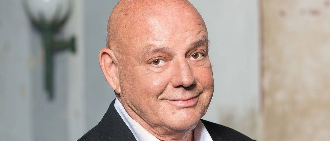 Ein älterer Mann mit kahlem Kopf und lächelndem Gesicht steht in einem Raum in einem schwarzen Anzug und einem weißen Hemd.