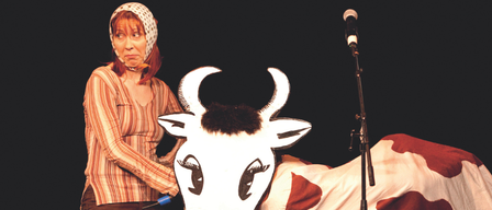 Eine Frau in einem gestreiften Shirt und einem weißen Kopftuch hält eine weiße Kuhkopfmaske mit einem Mikrofon dahinter.