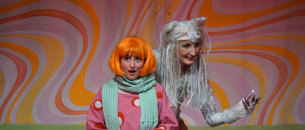 Zwei kostümierte Personen sind auf der Bühne, eine mit orangefarbenem Haar in einem rosa Outfit, die andere mit einem weißen Kostüm und einem Kopfschmuck. Sie sitzen auf einem grün drapierten Bett mit einem Hintergrund aus farbenfrohen Wirbeln.