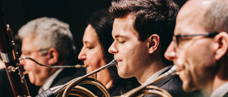 Vier Musiker in einer Reihe, gekleidet in formeller Kleidung, spielen französische Hörner. Der Mann in der Mitte konzentriert sich auf sein Instrument, während die anderen umherschauen.