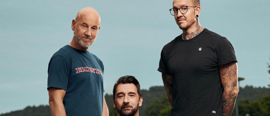 Drei Männer stehen auf einem Feld, zwei mit Hemden, einer mit Bart und Brille, der andere mit Tattoos und einer Uhr.