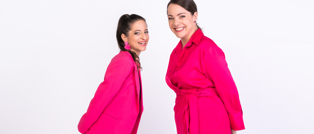 Zwei Frauen in leuchtend rosa Outfits lächeln und posieren für ein Foto vor weißem Hintergrund.