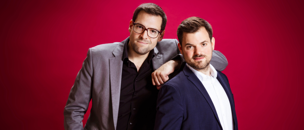 Zwei formell gekleidete Männer, einer mit Brille und der andere mit Bart, stehen vor einem roten Hintergrund.