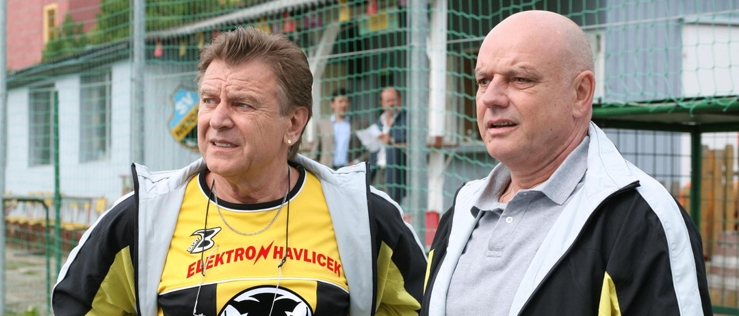 Zwei Männer stehen nebeneinander, einer trägt ein gelbes und schwarzes Sporttrikot mit einem Löwenemblem und dem Wort 'Elektro Havlicek'. Der andere Mann trägt ein graues Poloshirt. Hinter ihnen stehen zwei Personen in der Nähe eines Zauns.