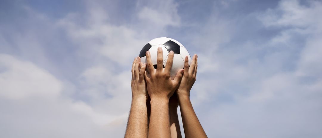 Drei Hände halten einen Fußball vor einem blauen Himmel mit verstreuten Wolken. Der Ball ist schwarz und weiß.