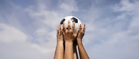 Drei Hände halten einen Fußball vor einem blauen Himmel mit verstreuten Wolken. Der Ball ist schwarz und weiß.