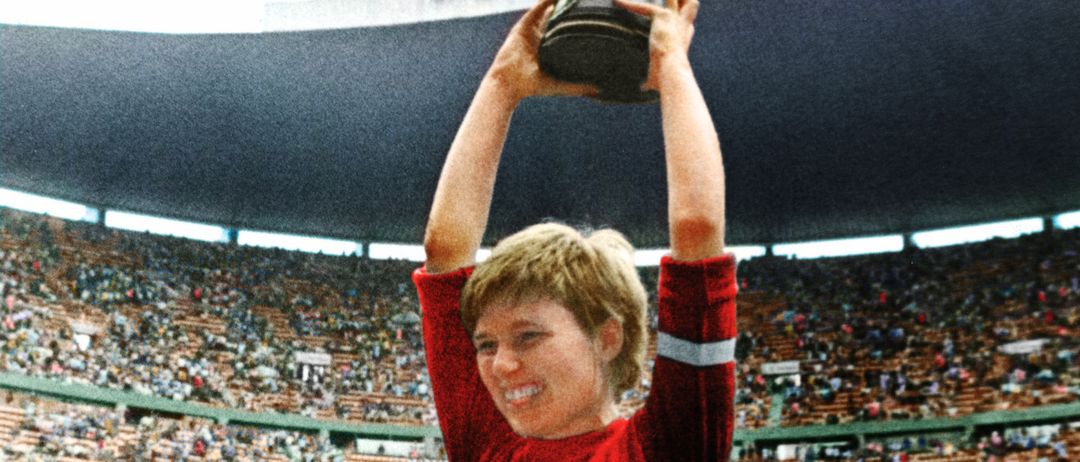 Ein junger Mann in einem roten Trikot wird von anderen hochgehoben, während er einen Pokal hält und lächelt, in einem vollen Stadion. Die Leute um ihn herum feiern.