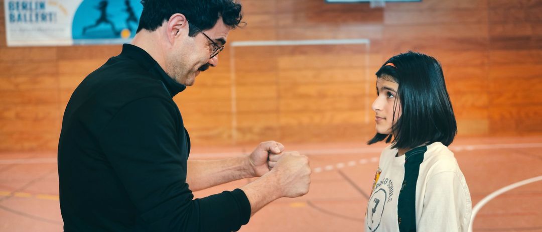 Ein Mann mit Brille und einem schwarzen Langarmshirt übt Handdrücken mit einem Mädchen in einem weißen Shirt in einer Turnhalle.